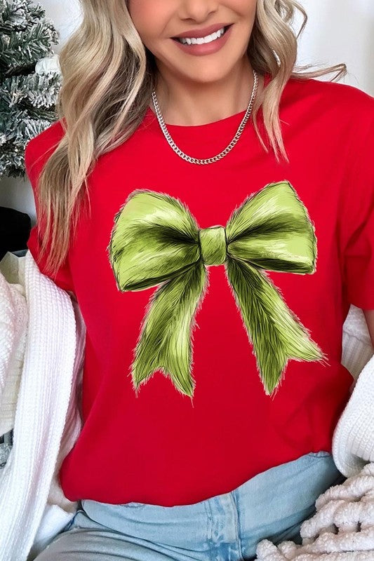 A person wearing a white Grinchmas Coquette Bow Graphic Tee with a green plaid shirt tied loosely over their shoulders. Only the lower part of their face is visible.