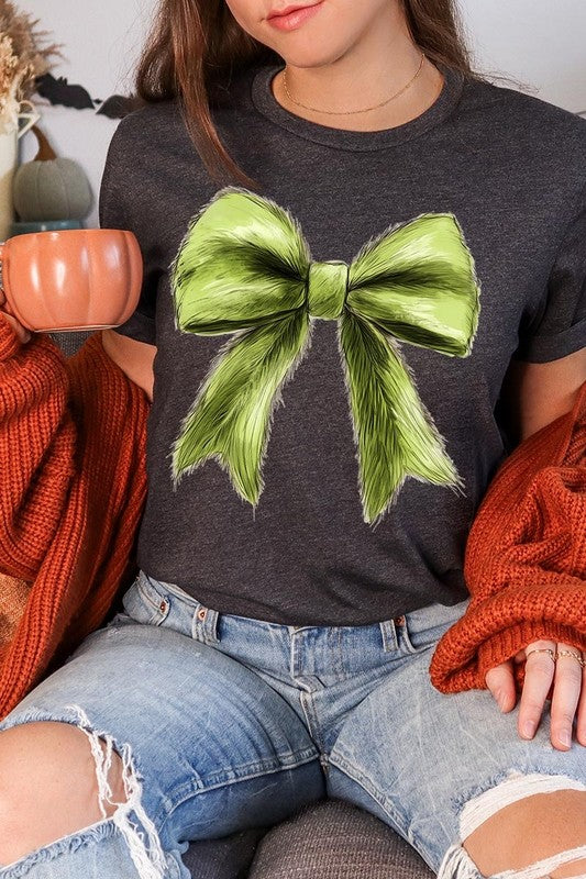 A person wearing a white Grinchmas Coquette Bow Graphic Tee with a green plaid shirt tied loosely over their shoulders. Only the lower part of their face is visible.