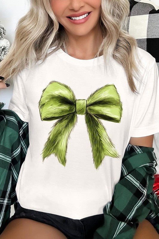 A person wearing a white Grinchmas Coquette Bow Graphic Tee with a green plaid shirt tied loosely over their shoulders. Only the lower part of their face is visible.