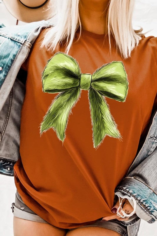 A person wearing a white Grinchmas Coquette Bow Graphic Tee with a green plaid shirt tied loosely over their shoulders. Only the lower part of their face is visible.