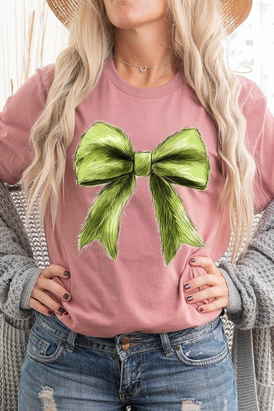 A person wearing a white Grinchmas Coquette Bow Graphic Tee with a green plaid shirt tied loosely over their shoulders. Only the lower part of their face is visible.