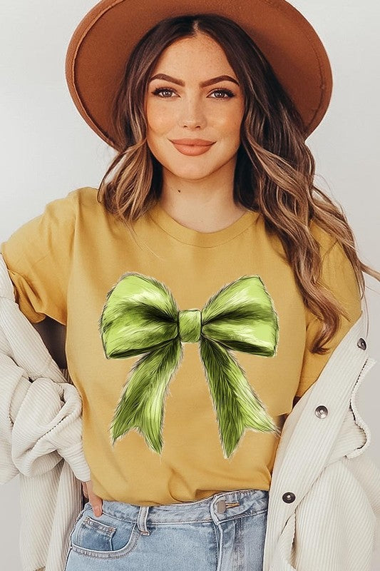 A person wearing a white Grinchmas Coquette Bow Graphic Tee with a green plaid shirt tied loosely over their shoulders. Only the lower part of their face is visible.