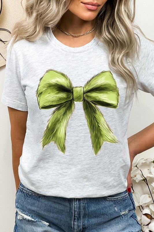 A person wearing a white Grinchmas Coquette Bow Graphic Tee with a green plaid shirt tied loosely over their shoulders. Only the lower part of their face is visible.