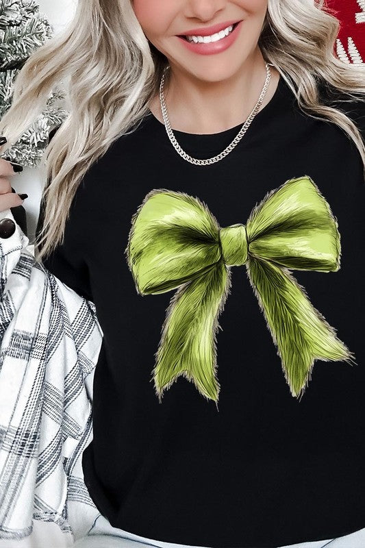 A person wearing a white Grinchmas Coquette Bow Graphic Tee with a green plaid shirt tied loosely over their shoulders. Only the lower part of their face is visible.