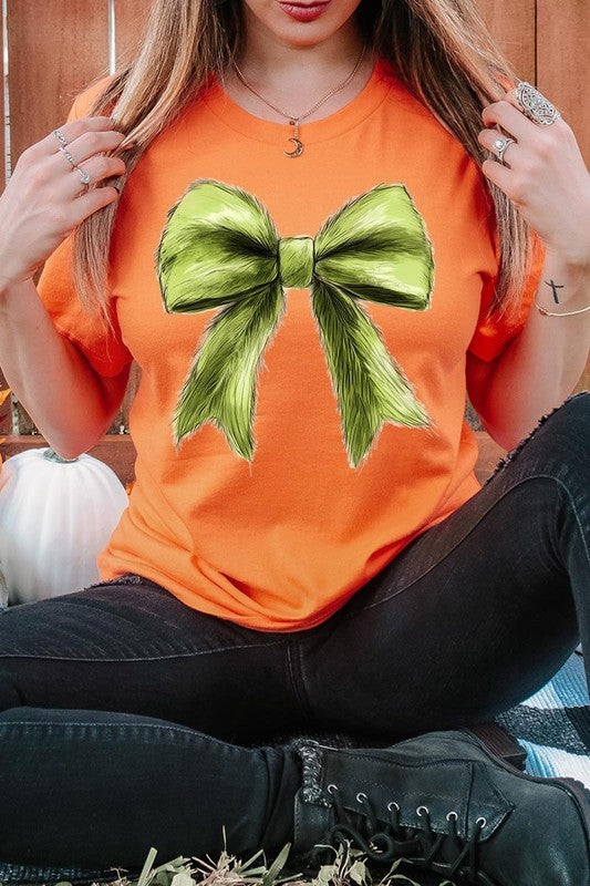 A person wearing a white Grinchmas Coquette Bow Graphic Tee with a green plaid shirt tied loosely over their shoulders. Only the lower part of their face is visible.