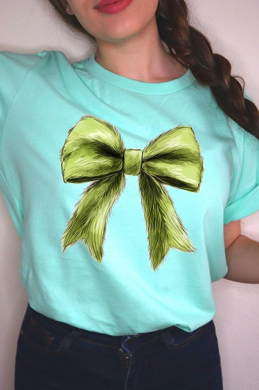 A person wearing a white Grinchmas Coquette Bow Graphic Tee with a green plaid shirt tied loosely over their shoulders. Only the lower part of their face is visible.