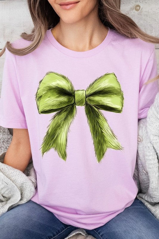 A person wearing a white Grinchmas Coquette Bow Graphic Tee with a green plaid shirt tied loosely over their shoulders. Only the lower part of their face is visible.