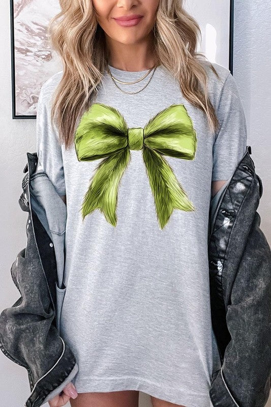 A person wearing a white Grinchmas Coquette Bow Graphic Tee with a green plaid shirt tied loosely over their shoulders. Only the lower part of their face is visible.