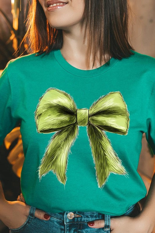 A person wearing a white Grinchmas Coquette Bow Graphic Tee with a green plaid shirt tied loosely over their shoulders. Only the lower part of their face is visible.