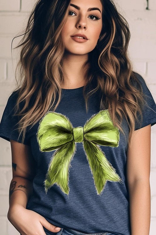 A person wearing a white Grinchmas Coquette Bow Graphic Tee with a green plaid shirt tied loosely over their shoulders. Only the lower part of their face is visible.