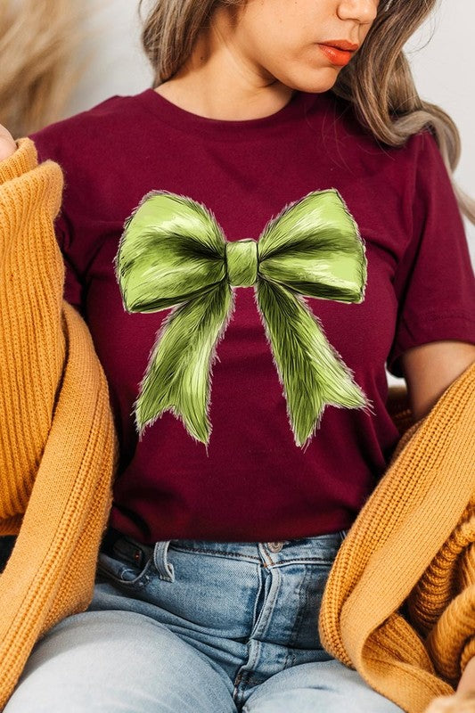 A person wearing a white Grinchmas Coquette Bow Graphic Tee with a green plaid shirt tied loosely over their shoulders. Only the lower part of their face is visible.