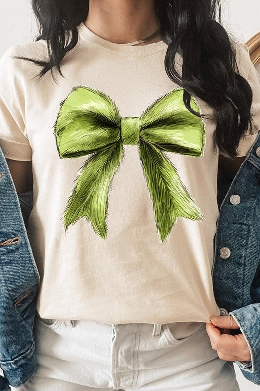 A person wearing a white Grinchmas Coquette Bow Graphic Tee with a green plaid shirt tied loosely over their shoulders. Only the lower part of their face is visible.