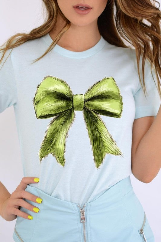 A person wearing a white Grinchmas Coquette Bow Graphic Tee with a green plaid shirt tied loosely over their shoulders. Only the lower part of their face is visible.