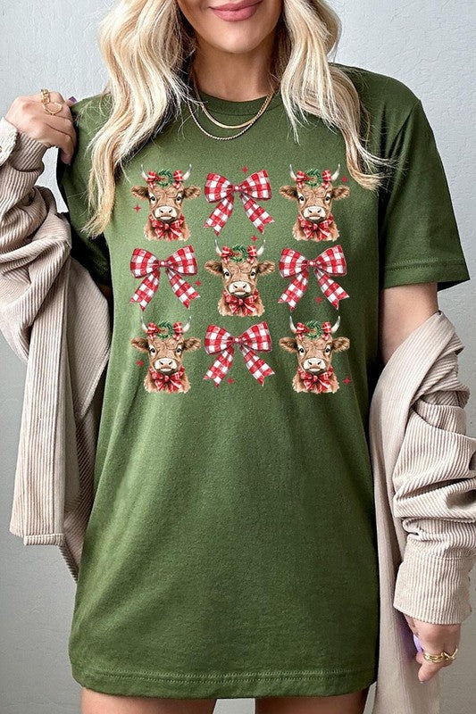 A person wearing a Christmas Highland Cow Graphic Tee in cream, showcasing six cartoon cows decorated with flowers and holly, alternated with red and white plaid bows. The person complements the look with a red and black plaid shirt. This unisex crew neck is ideal for family group uniforms during the festive season.