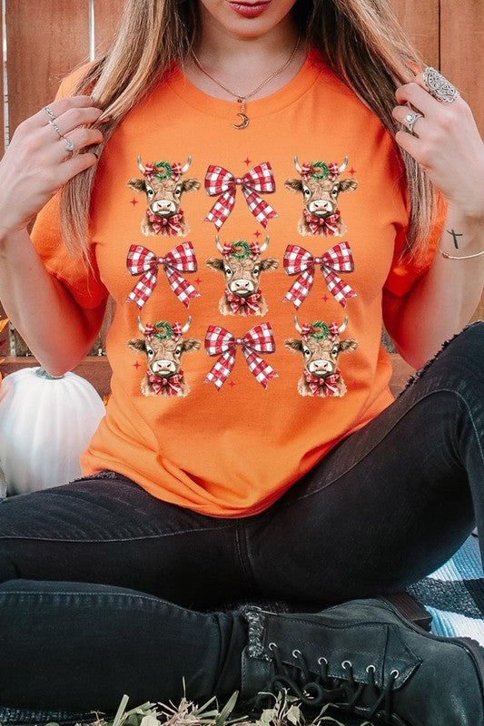 A person wearing a Christmas Highland Cow Graphic Tee in cream, showcasing six cartoon cows decorated with flowers and holly, alternated with red and white plaid bows. The person complements the look with a red and black plaid shirt. This unisex crew neck is ideal for family group uniforms during the festive season.