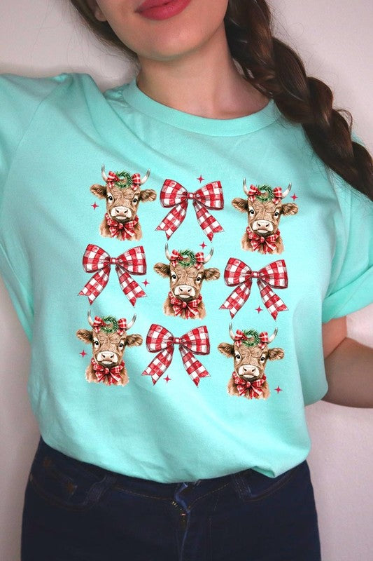 A person wearing a Christmas Highland Cow Graphic Tee in cream, showcasing six cartoon cows decorated with flowers and holly, alternated with red and white plaid bows. The person complements the look with a red and black plaid shirt. This unisex crew neck is ideal for family group uniforms during the festive season.