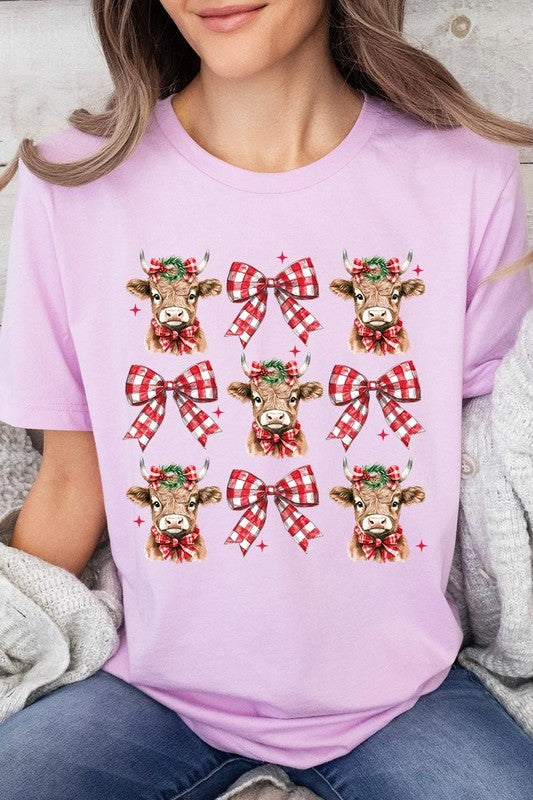 A person wearing a Christmas Highland Cow Graphic Tee in cream, showcasing six cartoon cows decorated with flowers and holly, alternated with red and white plaid bows. The person complements the look with a red and black plaid shirt. This unisex crew neck is ideal for family group uniforms during the festive season.