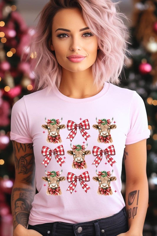 A person wearing a Christmas Highland Cow Graphic Tee in cream, showcasing six cartoon cows decorated with flowers and holly, alternated with red and white plaid bows. The person complements the look with a red and black plaid shirt. This unisex crew neck is ideal for family group uniforms during the festive season.