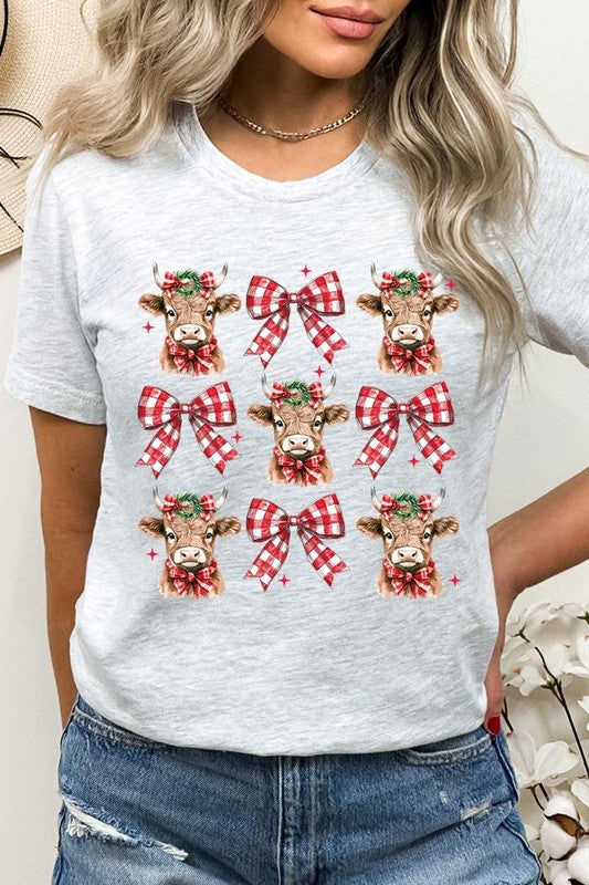 A person wearing a Christmas Highland Cow Graphic Tee in cream, showcasing six cartoon cows decorated with flowers and holly, alternated with red and white plaid bows. The person complements the look with a red and black plaid shirt. This unisex crew neck is ideal for family group uniforms during the festive season.