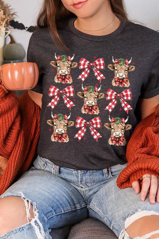 A person wearing a Christmas Highland Cow Graphic Tee in cream, showcasing six cartoon cows decorated with flowers and holly, alternated with red and white plaid bows. The person complements the look with a red and black plaid shirt. This unisex crew neck is ideal for family group uniforms during the festive season.