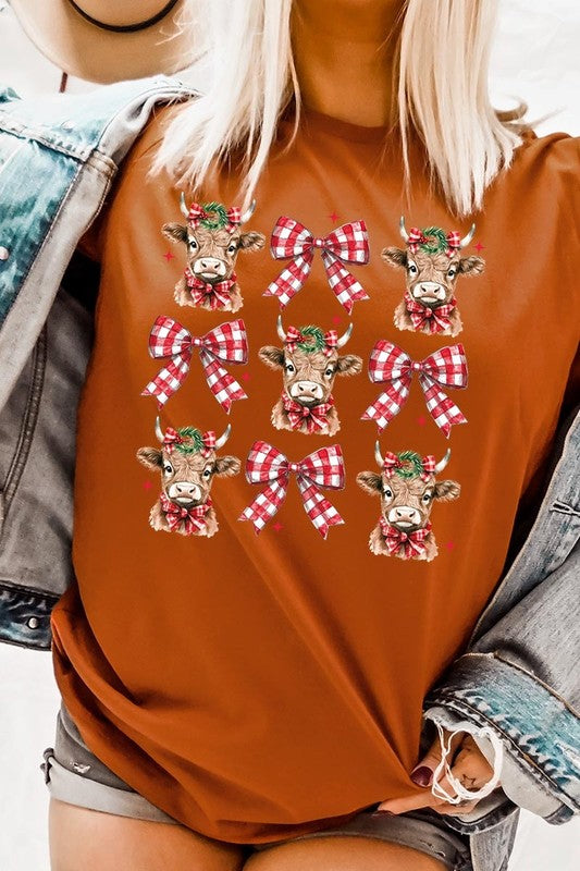 A person wearing a Christmas Highland Cow Graphic Tee in cream, showcasing six cartoon cows decorated with flowers and holly, alternated with red and white plaid bows. The person complements the look with a red and black plaid shirt. This unisex crew neck is ideal for family group uniforms during the festive season.