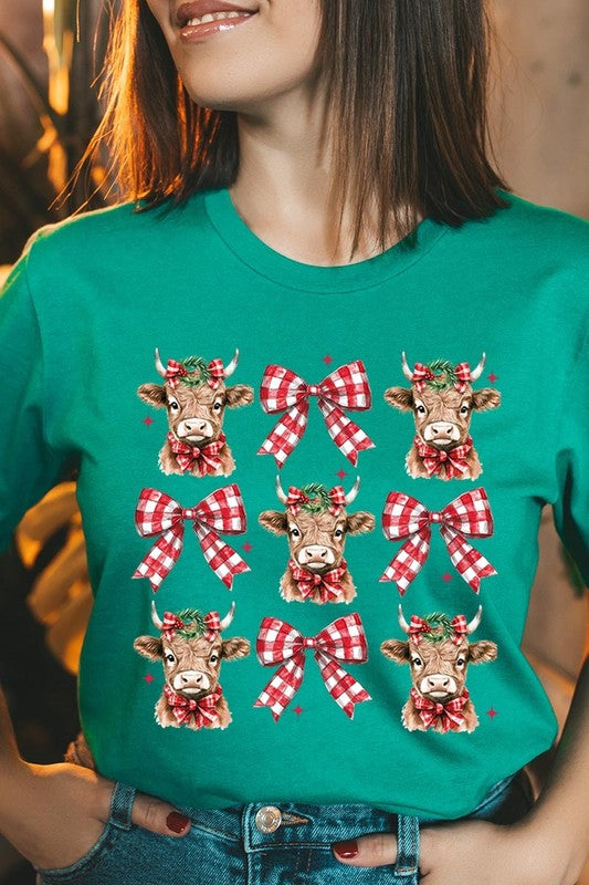 A person wearing a Christmas Highland Cow Graphic Tee in cream, showcasing six cartoon cows decorated with flowers and holly, alternated with red and white plaid bows. The person complements the look with a red and black plaid shirt. This unisex crew neck is ideal for family group uniforms during the festive season.