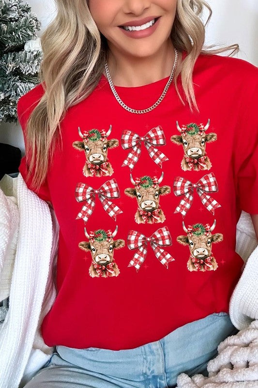 A person wearing a Christmas Highland Cow Graphic Tee in cream, showcasing six cartoon cows decorated with flowers and holly, alternated with red and white plaid bows. The person complements the look with a red and black plaid shirt. This unisex crew neck is ideal for family group uniforms during the festive season.