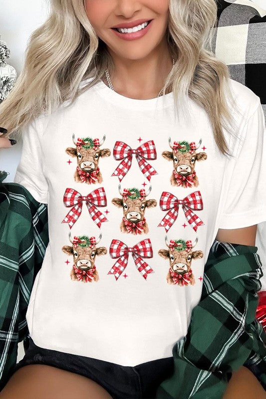 A person wearing a Christmas Highland Cow Graphic Tee in cream, showcasing six cartoon cows decorated with flowers and holly, alternated with red and white plaid bows. The person complements the look with a red and black plaid shirt. This unisex crew neck is ideal for family group uniforms during the festive season.