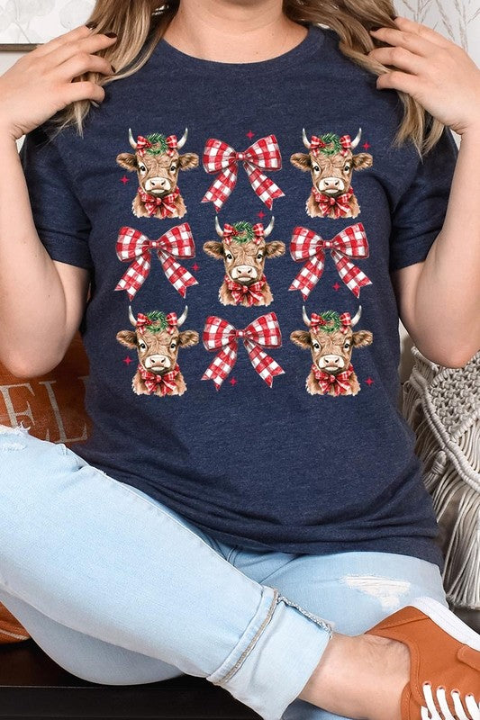 A person wearing a Christmas Highland Cow Graphic Tee in cream, showcasing six cartoon cows decorated with flowers and holly, alternated with red and white plaid bows. The person complements the look with a red and black plaid shirt. This unisex crew neck is ideal for family group uniforms during the festive season.