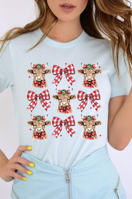 A person wearing a Christmas Highland Cow Graphic Tee in cream, showcasing six cartoon cows decorated with flowers and holly, alternated with red and white plaid bows. The person complements the look with a red and black plaid shirt. This unisex crew neck is ideal for family group uniforms during the festive season.