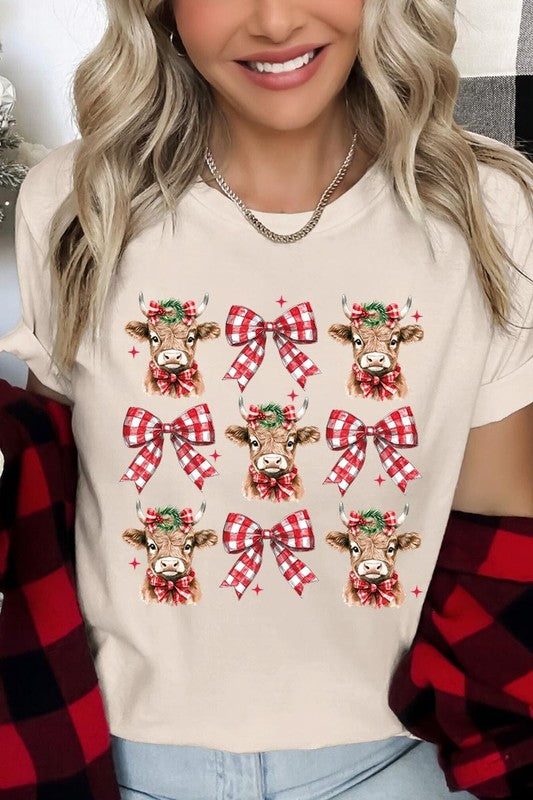 A person wearing a Christmas Highland Cow Graphic Tee in cream, showcasing six cartoon cows decorated with flowers and holly, alternated with red and white plaid bows. The person complements the look with a red and black plaid shirt. This unisex crew neck is ideal for family group uniforms during the festive season.