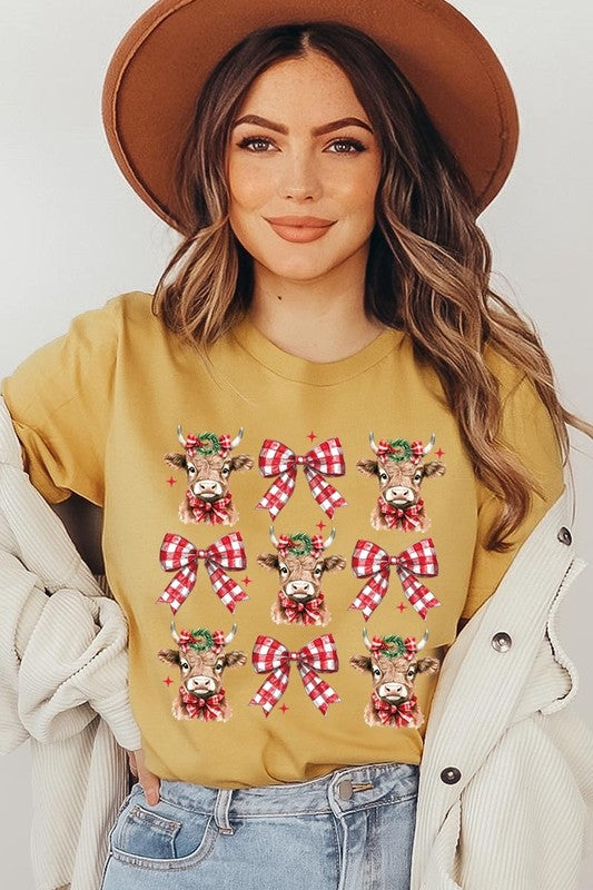 A person wearing a Christmas Highland Cow Graphic Tee in cream, showcasing six cartoon cows decorated with flowers and holly, alternated with red and white plaid bows. The person complements the look with a red and black plaid shirt. This unisex crew neck is ideal for family group uniforms during the festive season.