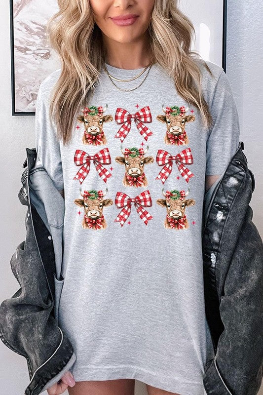 A person wearing a Christmas Highland Cow Graphic Tee in cream, showcasing six cartoon cows decorated with flowers and holly, alternated with red and white plaid bows. The person complements the look with a red and black plaid shirt. This unisex crew neck is ideal for family group uniforms during the festive season.