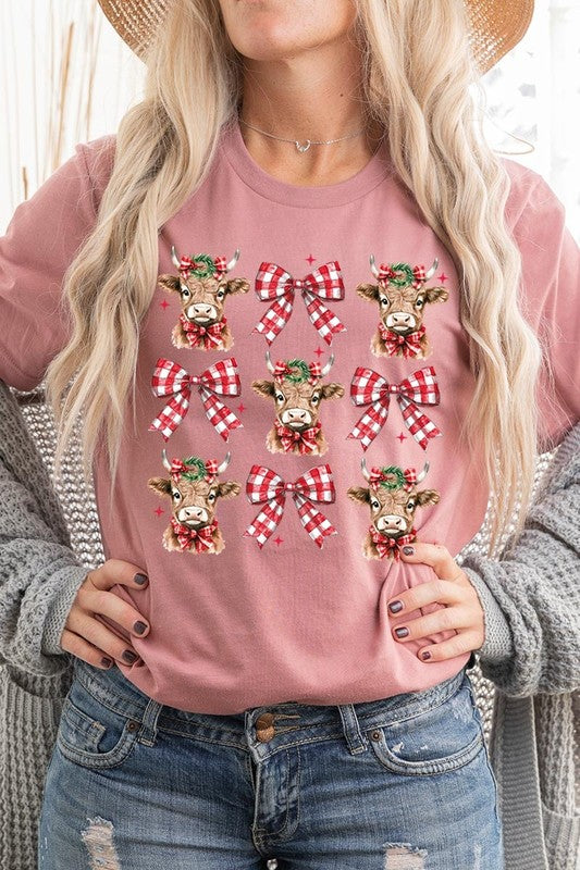 A person wearing a Christmas Highland Cow Graphic Tee in cream, showcasing six cartoon cows decorated with flowers and holly, alternated with red and white plaid bows. The person complements the look with a red and black plaid shirt. This unisex crew neck is ideal for family group uniforms during the festive season.