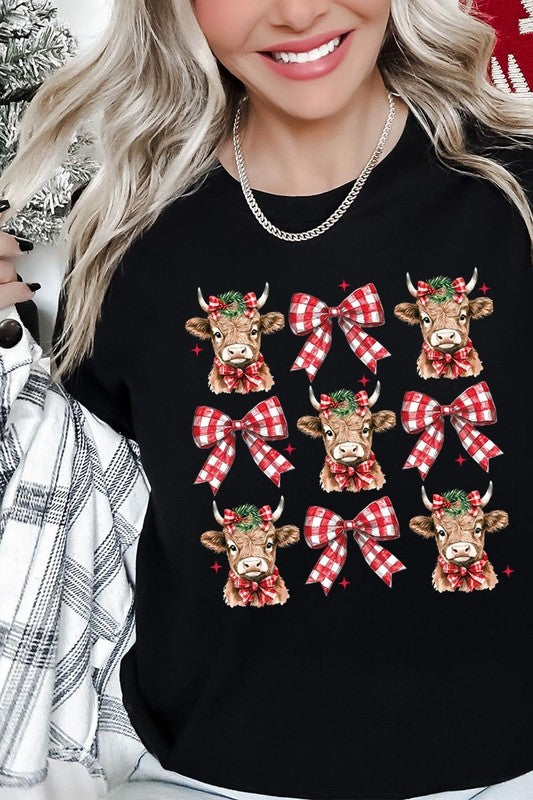 A person wearing a Christmas Highland Cow Graphic Tee in cream, showcasing six cartoon cows decorated with flowers and holly, alternated with red and white plaid bows. The person complements the look with a red and black plaid shirt. This unisex crew neck is ideal for family group uniforms during the festive season.