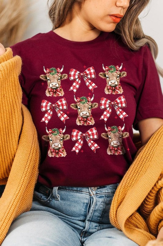 A person wearing a Christmas Highland Cow Graphic Tee in cream, showcasing six cartoon cows decorated with flowers and holly, alternated with red and white plaid bows. The person complements the look with a red and black plaid shirt. This unisex crew neck is ideal for family group uniforms during the festive season.