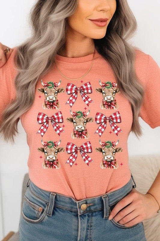 A person wearing a Christmas Highland Cow Graphic Tee in cream, showcasing six cartoon cows decorated with flowers and holly, alternated with red and white plaid bows. The person complements the look with a red and black plaid shirt. This unisex crew neck is ideal for family group uniforms during the festive season.