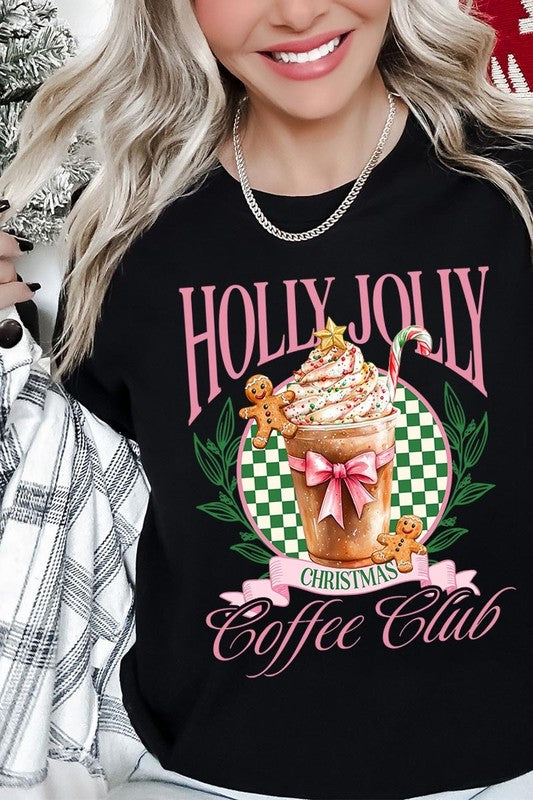 A woman with pink hair is wearing the "Holly Jolly Christmas Coffee Club Graphic Tee," adorned with a charming design of a festive coffee cup and gingerbread men. The Christmas decorations in the background enhance the jolly atmosphere of this unisex crew neck shirt.