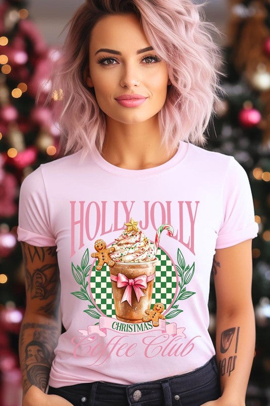 A woman with pink hair is wearing the "Holly Jolly Christmas Coffee Club Graphic Tee," adorned with a charming design of a festive coffee cup and gingerbread men. The Christmas decorations in the background enhance the jolly atmosphere of this unisex crew neck shirt.