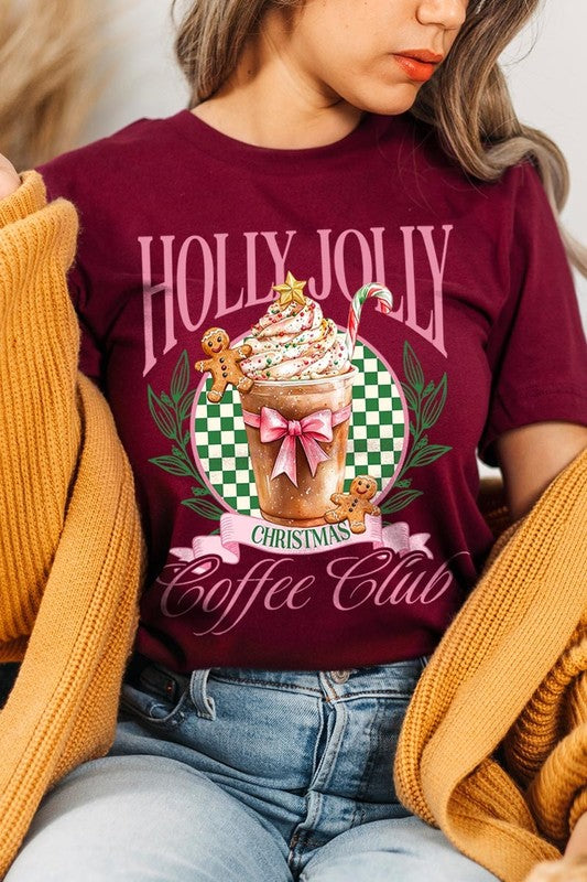 A woman with pink hair is wearing the "Holly Jolly Christmas Coffee Club Graphic Tee," adorned with a charming design of a festive coffee cup and gingerbread men. The Christmas decorations in the background enhance the jolly atmosphere of this unisex crew neck shirt.