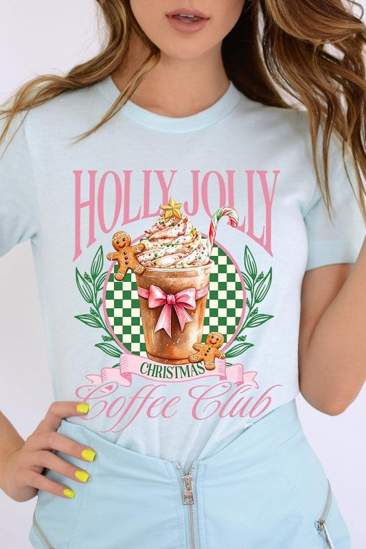 A woman with pink hair is wearing the "Holly Jolly Christmas Coffee Club Graphic Tee," adorned with a charming design of a festive coffee cup and gingerbread men. The Christmas decorations in the background enhance the jolly atmosphere of this unisex crew neck shirt.