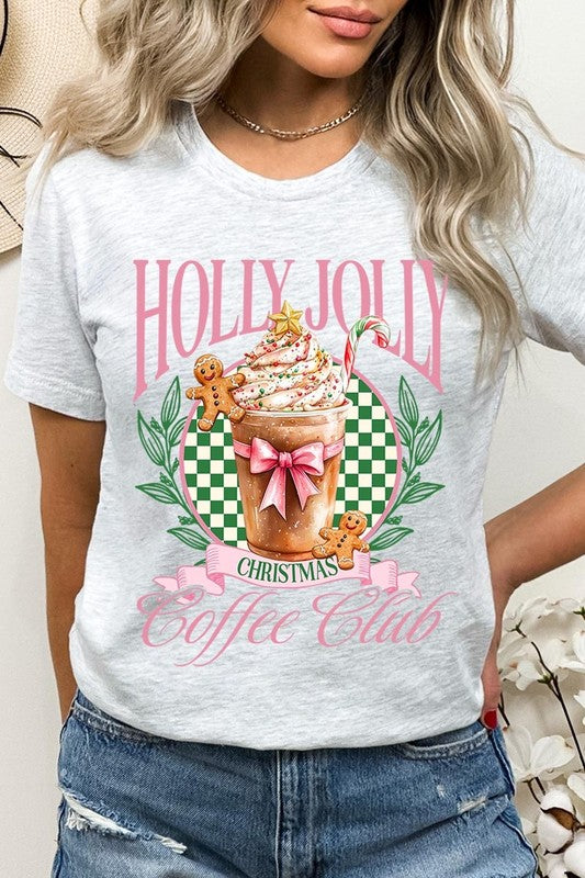 A woman with pink hair is wearing the "Holly Jolly Christmas Coffee Club Graphic Tee," adorned with a charming design of a festive coffee cup and gingerbread men. The Christmas decorations in the background enhance the jolly atmosphere of this unisex crew neck shirt.