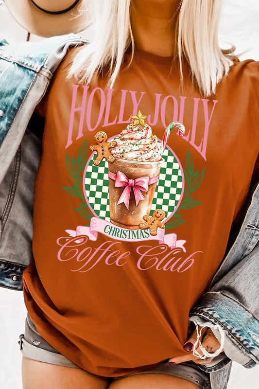 A woman with pink hair is wearing the "Holly Jolly Christmas Coffee Club Graphic Tee," adorned with a charming design of a festive coffee cup and gingerbread men. The Christmas decorations in the background enhance the jolly atmosphere of this unisex crew neck shirt.