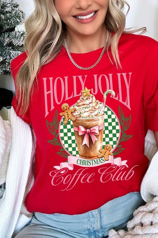 A woman with pink hair is wearing the "Holly Jolly Christmas Coffee Club Graphic Tee," adorned with a charming design of a festive coffee cup and gingerbread men. The Christmas decorations in the background enhance the jolly atmosphere of this unisex crew neck shirt.