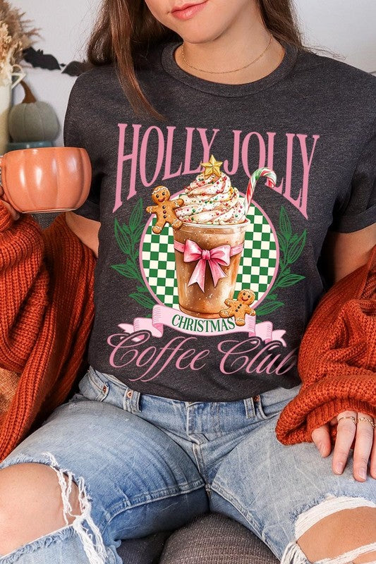 A woman with pink hair is wearing the "Holly Jolly Christmas Coffee Club Graphic Tee," adorned with a charming design of a festive coffee cup and gingerbread men. The Christmas decorations in the background enhance the jolly atmosphere of this unisex crew neck shirt.