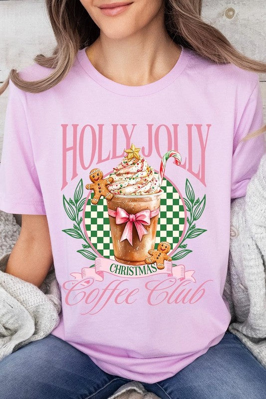 A woman with pink hair is wearing the "Holly Jolly Christmas Coffee Club Graphic Tee," adorned with a charming design of a festive coffee cup and gingerbread men. The Christmas decorations in the background enhance the jolly atmosphere of this unisex crew neck shirt.