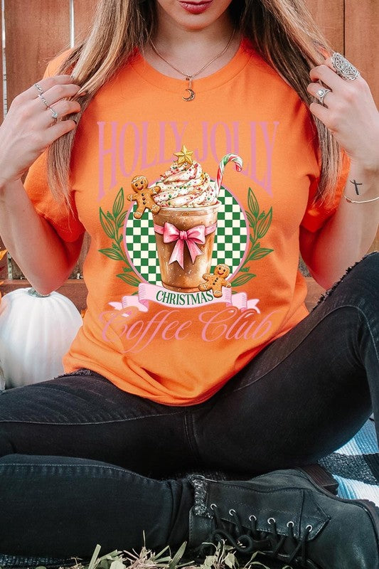 A woman with pink hair is wearing the "Holly Jolly Christmas Coffee Club Graphic Tee," adorned with a charming design of a festive coffee cup and gingerbread men. The Christmas decorations in the background enhance the jolly atmosphere of this unisex crew neck shirt.