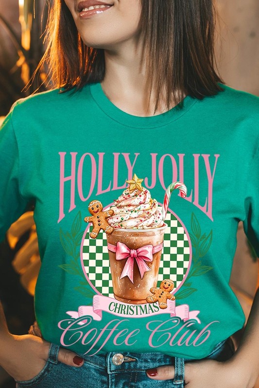 A woman with pink hair is wearing the "Holly Jolly Christmas Coffee Club Graphic Tee," adorned with a charming design of a festive coffee cup and gingerbread men. The Christmas decorations in the background enhance the jolly atmosphere of this unisex crew neck shirt.