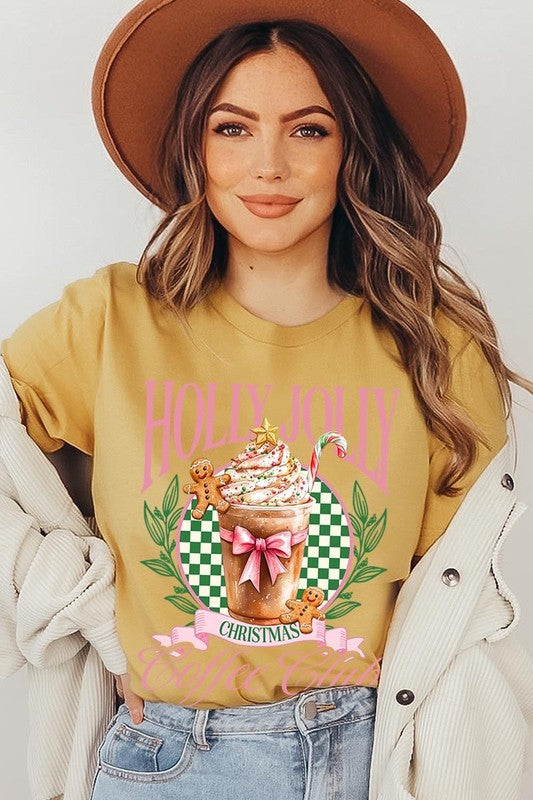 A woman with pink hair is wearing the "Holly Jolly Christmas Coffee Club Graphic Tee," adorned with a charming design of a festive coffee cup and gingerbread men. The Christmas decorations in the background enhance the jolly atmosphere of this unisex crew neck shirt.