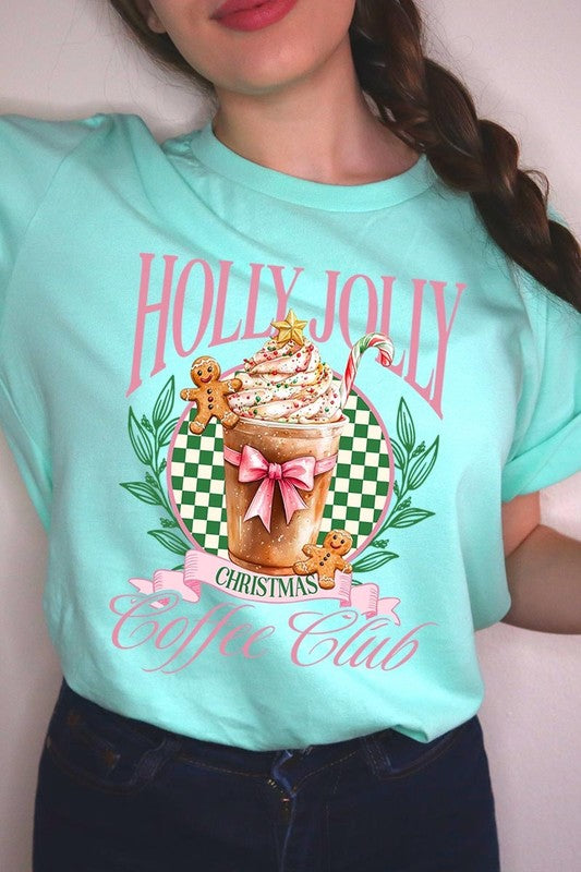 A woman with pink hair is wearing the "Holly Jolly Christmas Coffee Club Graphic Tee," adorned with a charming design of a festive coffee cup and gingerbread men. The Christmas decorations in the background enhance the jolly atmosphere of this unisex crew neck shirt.